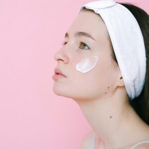 A serene woman applying skincare cream on her cheek against a pink background.