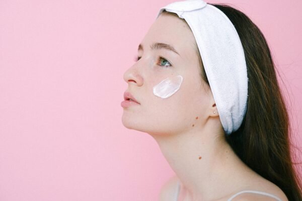 A serene woman applying skincare cream on her cheek against a pink background.