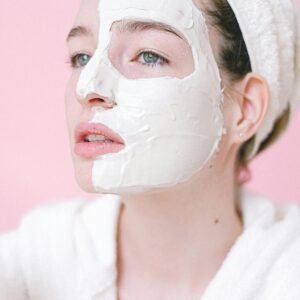 Serene female in bath robe and towel on head standing with applied facial mask on half face in light spa salon and looking away