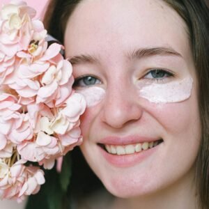 Crop positive young female model with eyes mask smiling at camera while demonstrating hydrangea flower on pink background