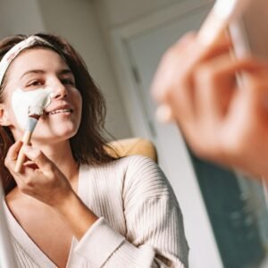 Woman enjoys a skincare routine, applying a facial mask with a brush in front of a mirror.