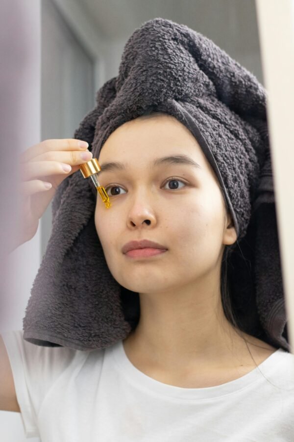 An Asian woman applies skincare serum with a dropper in front of a mirror.