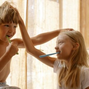 Two cheerful children practicing oral hygiene together, brushing teeth in a sunlit bathroom setting.