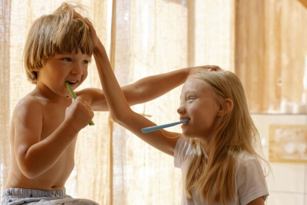 Two cheerful children practicing oral hygiene together, brushing teeth in a sunlit bathroom setting.