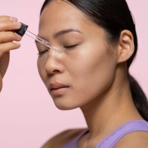 Close-up of a woman applying facial serum using a dropper on a pink background.