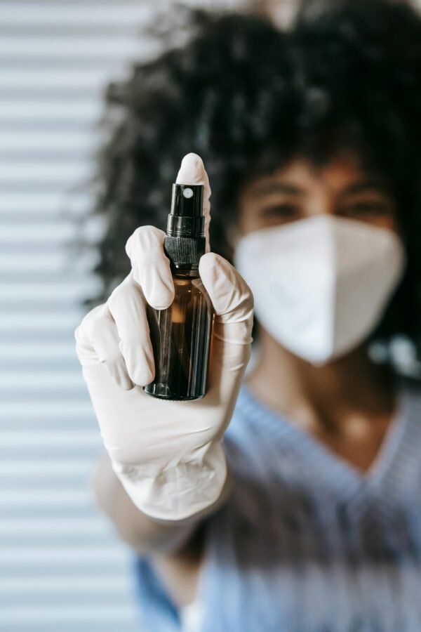 Blurred African American female wearing white protective mask and latex medical sterile gloves spraying antiseptic in room and looking at camera